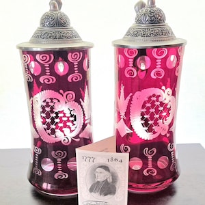 May include: Two ruby red glass beer steins with ornate silver lids. The steins feature etched floral designs and decorative cutouts. A small card with a portrait and the dates 1777 and 1864 is in front of the steins.