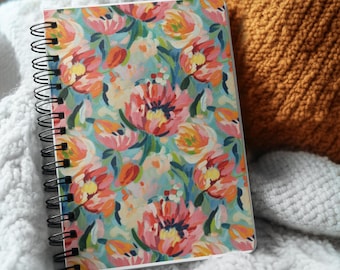 May include: A spiral-bound notebook with a floral design. The cover features a pattern of pink, orange, and yellow flowers on a light blue background. The notebook is resting on a white, textured blanket, with a mustard-colored pillow in the background.