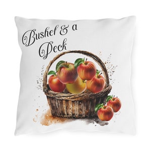 May include: A white pillow with the text "Bushel & a Peck" in black cursive font. A brown wicker basket filled with red apples and a yellow pear is in the center of the pillow. The background is a watercolor wash of white, brown, and orange.