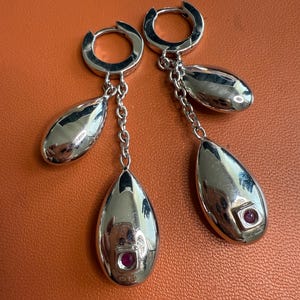May include: A pair of silver-toned earrings with a teardrop design. Each earring features a small, round, red gemstone set in a square bezel. The earrings hang from a chain and a hoop closure.