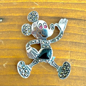 May include: Silver-toned Mickey Mouse brooch with marcasite detailing. The brooch features a black square for the shorts, pink gems for the eyes, and is posed with one arm raised. The brooch is on a wooden surface.