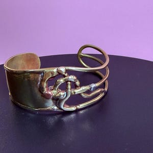 May include: A gold-toned cuff bracelet with an abstract design. The bracelet features a wide band and an intricate overlay of curved and looped metal elements. The design incorporates silver and copper accents, creating a unique, artistic piece of jewelry.