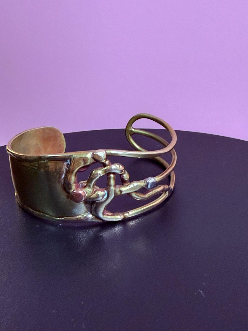 May include: A gold-toned cuff bracelet with an abstract design. The bracelet features a wide band and an intricate overlay of curved and looped metal elements. The design incorporates silver and copper accents, creating a unique, artistic piece of jewelry.