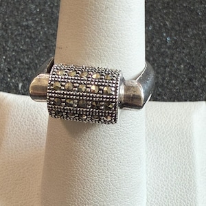 May include: A silver ring with a rectangular center section encrusted with small, sparkling stones. The ring is displayed on a white surface, showcasing its design and details. The band has a simple, polished finish.