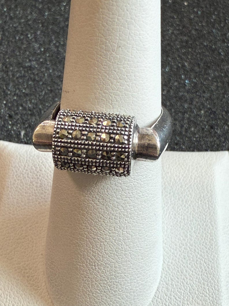 May include: A silver ring with a rectangular center section encrusted with small, sparkling stones. The ring is displayed on a white surface, showcasing its design and details. The band has a simple, polished finish.