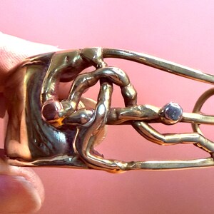 May include: A gold-toned metal bracelet with an abstract design. The bracelet features intertwined shapes and a small, round, gray stone. The design has a textured appearance and is set against a pink background.