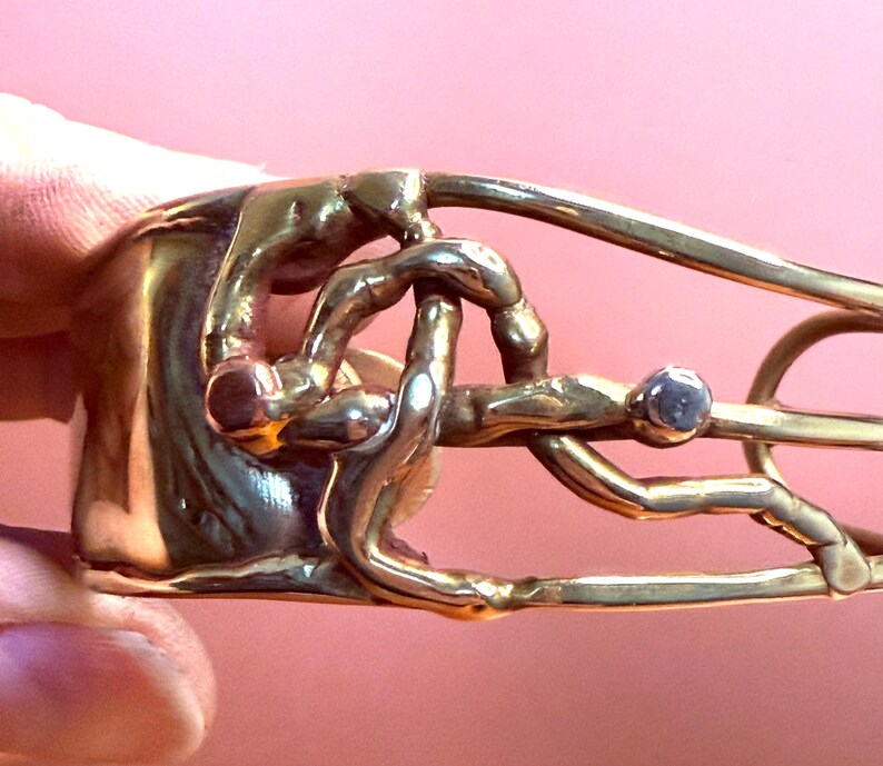 May include: A gold-toned metal bracelet with an abstract design. The bracelet features intertwined shapes and a small, round, gray stone. The design has a textured appearance and is set against a pink background.