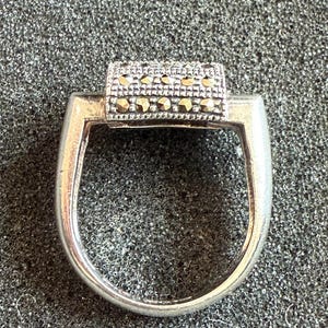 May include: A silver ring with a rectangular top adorned with small, sparkling stones. The ring has a simple, elegant design, perfect for everyday wear. The stones are set in a row, adding a touch of glamour to the piece.
