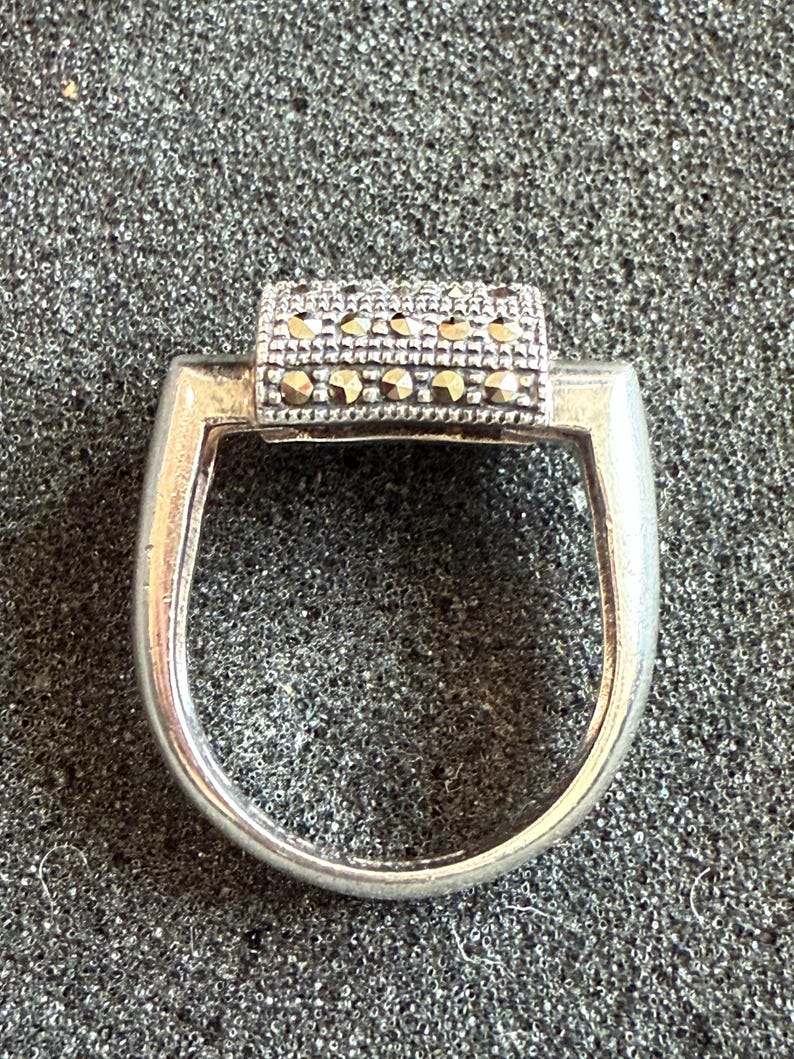 May include: A silver ring with a rectangular top adorned with small, sparkling stones. The ring has a simple, elegant design, perfect for everyday wear. The stones are set in a row, adding a touch of glamour to the piece.