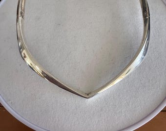 Könnte beinhalten: Eine silberfarbene Halskette mit V-förmiger Vorderseite. Die Halskette hat ein schlichtes, elegantes Design und einen Verschluss. Der Schmuck wird auf einer weißen Oberfläche präsentiert.