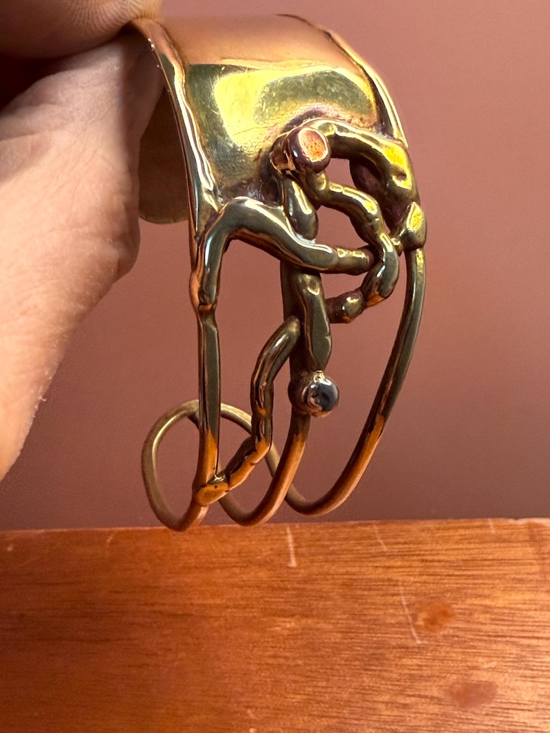 May include: A gold-toned metal cuff bracelet with an abstract design. The bracelet features a series of interconnected, organic shapes and a small, dark-colored stone. The cuff is held by a hand, showcasing its intricate details and craftsmanship.