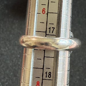 May include: A silver ring is placed on a ring sizer. The ring is a size 6, as indicated by the sizer. The ring is a simple band design, with a smooth, rounded surface. The sizer has black and white markings.