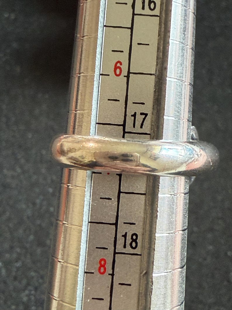 May include: A silver ring is placed on a ring sizer. The ring is a size 6, as indicated by the sizer. The ring is a simple band design, with a smooth, rounded surface. The sizer has black and white markings.