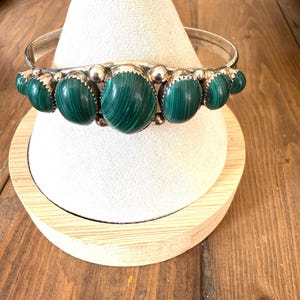 May include: A silver bangle bracelet with oval, vivid green malachite stones. The stones are set in a silver frame and are evenly spaced around the bracelet. The bracelet is displayed on a white, cone-shaped stand.
