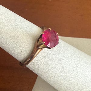 May include: A gold-toned ring featuring a large, faceted, oval-shaped ruby gemstone. The ring is set on a white surface, showcasing the intricate details of the setting and the vibrant red color of the gemstone.