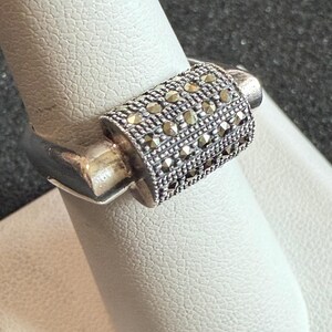 May include: A silver ring with a rectangular, textured band encrusted with small, round, dark-colored stones. The ring is displayed on a white surface, showcasing its design and craftsmanship. The ring's band is approximately 0.5 inches wide.