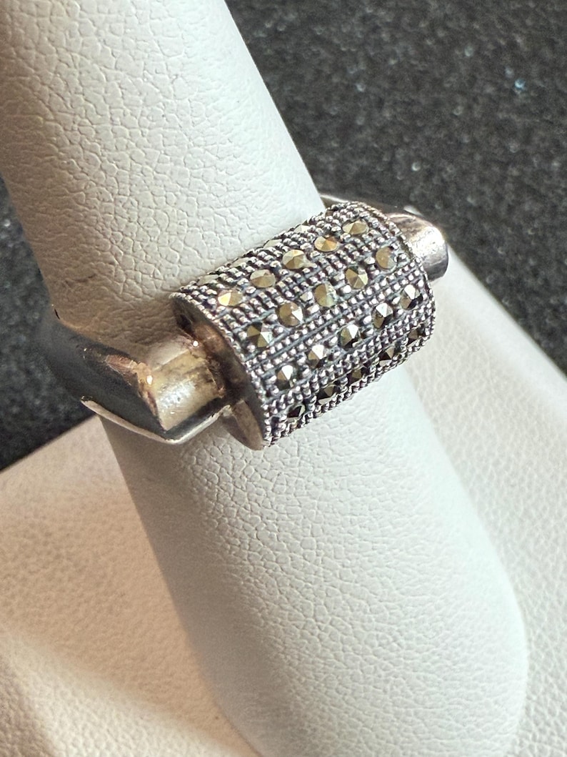 May include: A silver ring with a rectangular, textured band encrusted with small, round, dark-colored stones. The ring is displayed on a white surface, showcasing its design and craftsmanship. The ring's band is approximately 0.5 inches wide.