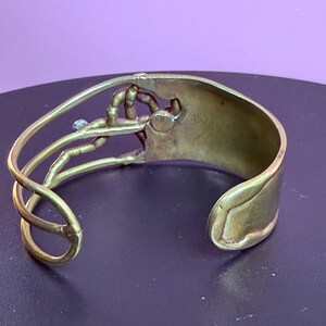 May include: A brass cuff bracelet with an open design. The bracelet features a stylized hand holding a small, round object. The cuff has a curved shape and a smooth, polished finish. A small, blue stone is set near the hand.