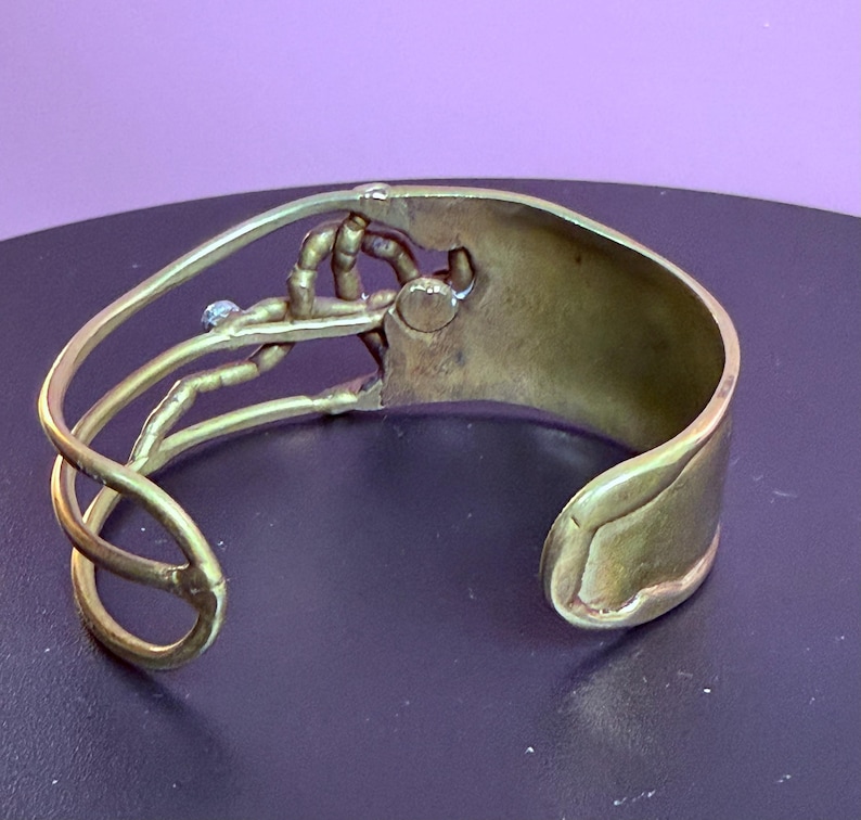 May include: A brass cuff bracelet with an open design. The bracelet features a stylized hand holding a small, round object. The cuff has a curved shape and a smooth, polished finish. A small, blue stone is set near the hand.