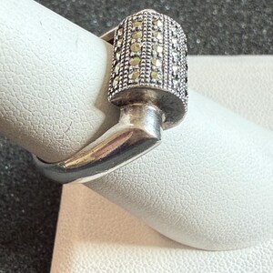 May include: A silver ring with a rectangular top encrusted with small, sparkling stones. The ring's design features a unique, angular shape, with a band that curves around the finger. The ring is displayed on a white surface.
