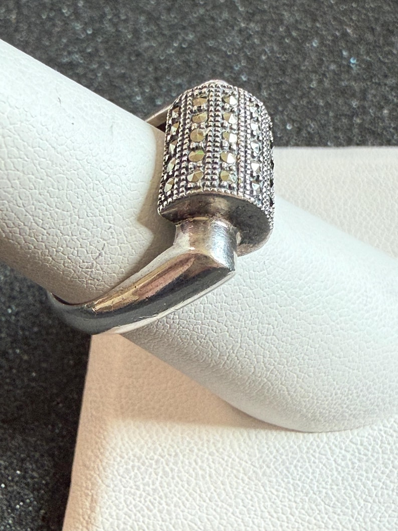 May include: A silver ring with a rectangular top encrusted with small, sparkling stones. The ring's design features a unique, angular shape, with a band that curves around the finger. The ring is displayed on a white surface.