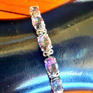 May include: A silver bracelet with oval, iridescent gemstones. The stones show purple, green, and blue hues. The bracelet has a delicate, ornate design, with silver links connecting the stones.