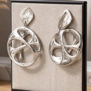May include: A pair of silver-toned, abstract earrings. Each earring features a leaf-shaped top connected to a large, openwork circular design with an interwoven pattern. The earrings are displayed against a neutral background.