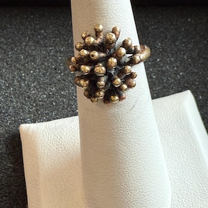 May include: A bronze-toned ring with a cluster design, resembling a coral formation. The ring features numerous small, rounded protrusions in a gold color, set against a darker, textured base. The ring is displayed on a white finger form.