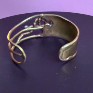 May include: A gold-toned cuff bracelet with an open design. The bracelet features a wide band and an intricate, decorative element on the top. The decorative element has a floral or organic shape. The cuff is likely made of metal.