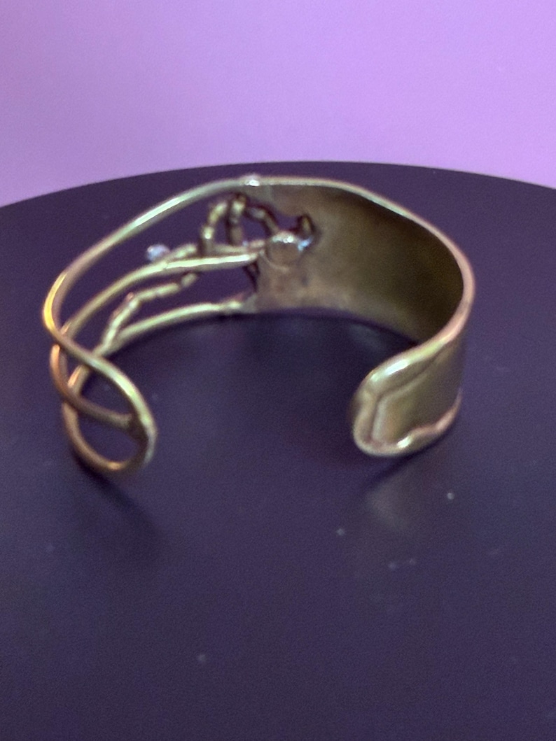 May include: A gold-toned cuff bracelet with an open design. The bracelet features a wide band and an intricate, decorative element on the top. The decorative element has a floral or organic shape. The cuff is likely made of metal.