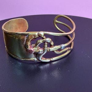 May include: A brass cuff bracelet with an abstract design. The bracelet features a wide band and a series of intertwined metal elements. The metal has a warm, golden hue with some areas showing darker oxidation. The design includes a small, silver-colored accent.