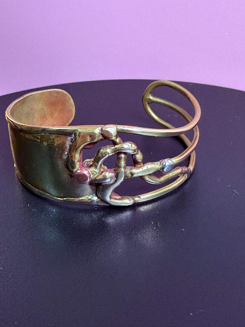 May include: A brass cuff bracelet with an abstract design. The bracelet features a wide band and a series of intertwined metal elements. The metal has a warm, golden hue with some areas showing darker oxidation. The design includes a small, silver-colored accent.