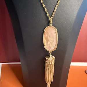 May include: Gold-toned necklace with a large, oval, light pink stone pendant. The pendant is framed in gold and has a decorative tassel of gold chains. The necklace is displayed on a black mannequin.