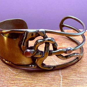 May include: A vintage brass cuff bracelet with an open design. The bracelet features a wide band and an intricate, abstract overlay with a small, red accent. The metal has a warm, aged patina, suggesting it is a handcrafted piece.