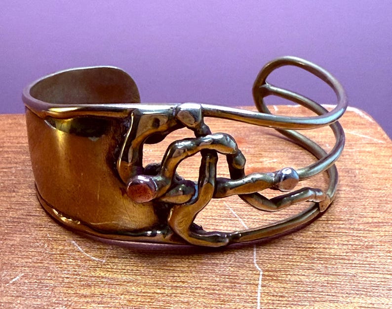 May include: A vintage brass cuff bracelet with an open design. The bracelet features a wide band and an intricate, abstract overlay with a small, red accent. The metal has a warm, aged patina, suggesting it is a handcrafted piece.