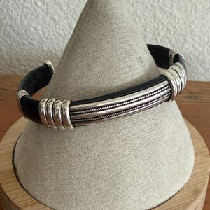 May include: A black leather cuff bracelet with silver-colored metal accents. The bracelet features a central band of twisted metal and is displayed on a gray, cone-shaped stand.