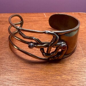 May include: A copper-toned metal cuff bracelet with an open design. The bracelet features a wide band and an intricate, sculptural overlay with a small, round embellishment. The metal has a warm, antique finish, and the design is abstract and artistic.