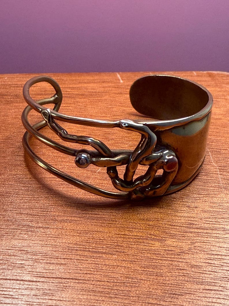 May include: A copper-toned metal cuff bracelet with an open design. The bracelet features a wide band and an intricate, sculptural overlay with a small, round embellishment. The metal has a warm, antique finish, and the design is abstract and artistic.