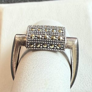 May include: A silver ring with a rectangular, marcasite-studded center. The ring's design features a barrel-shaped centerpiece with small, sparkling stones, and a curved band that extends to the sides.