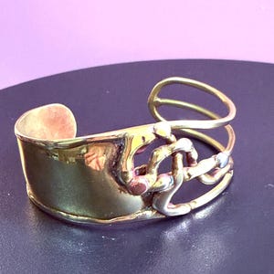 May include: A handcrafted brass cuff bracelet with a unique, abstract design. The bracelet features a wide, curved band with a textured surface and an open, looping design on the side. Accents include copper and silver-toned metal.