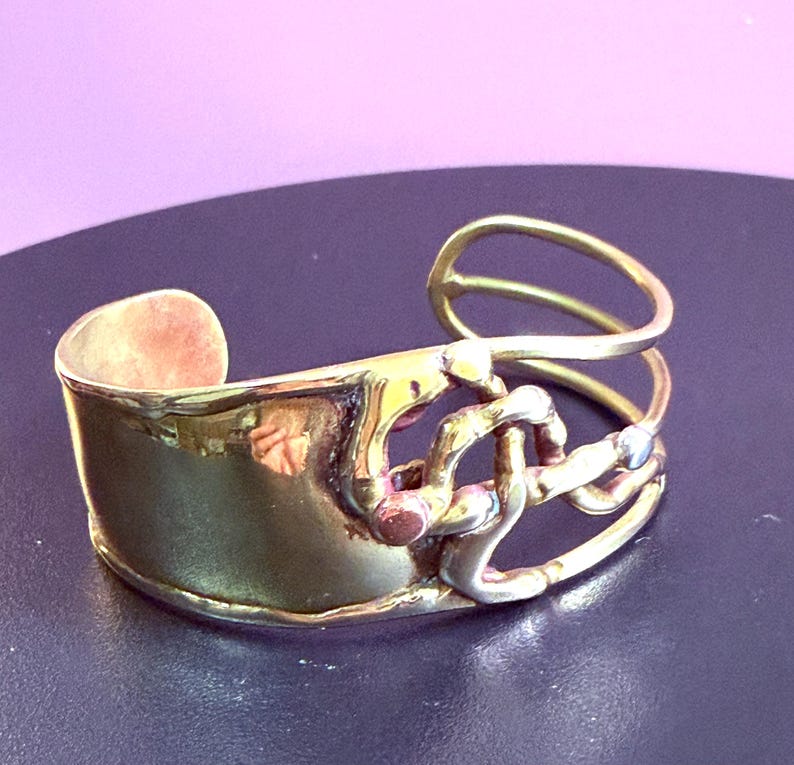 May include: A handcrafted brass cuff bracelet with a unique, abstract design. The bracelet features a wide, curved band with a textured surface and an open, looping design on the side. Accents include copper and silver-toned metal.