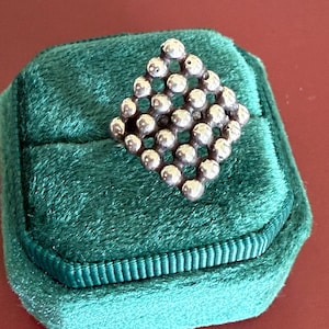 May include: A silver-toned ring with a geometric design of small spheres, set on a dark green velvet ring box. The ring's design is a diamond shape made of connected spheres. The ring box has a ribbed border.