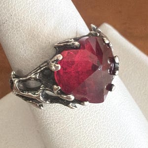 May include: A silver-toned ring featuring a large, faceted, deep red gemstone. The ring has an ornate, textured setting with a dark, oxidized finish. The gemstone is the focal point, catching the light and displaying a vibrant color.