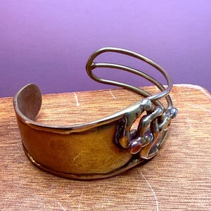 May include: A brass cuff bracelet with a wide band and an open design. The bracelet features a decorative element with three curved bars and small, colorful stones. The metal has a warm, golden hue.