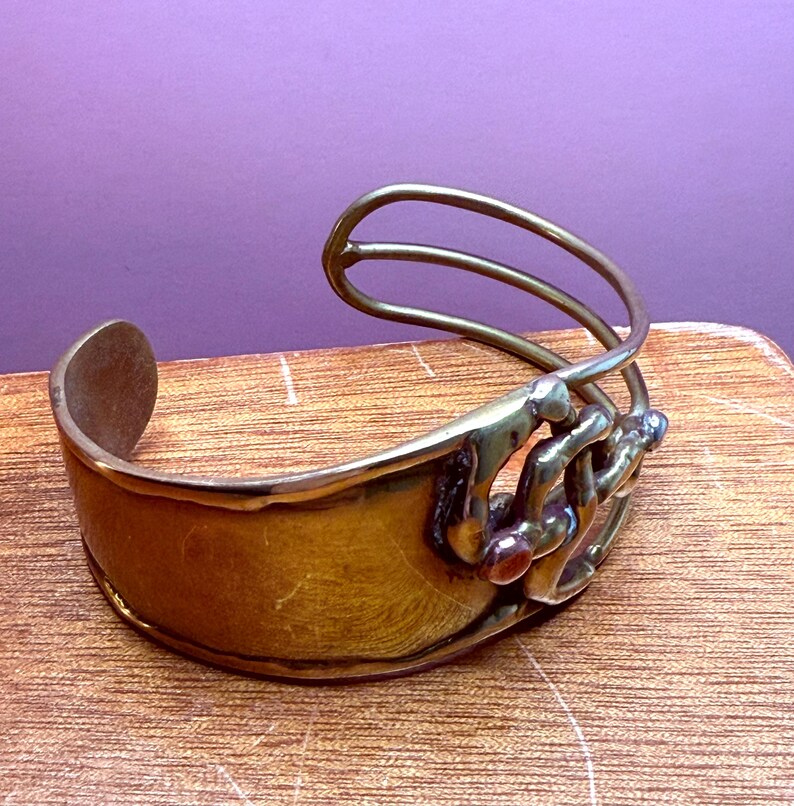 May include: A brass cuff bracelet with a wide band and an open design. The bracelet features a decorative element with three curved bars and small, colorful stones. The metal has a warm, golden hue.