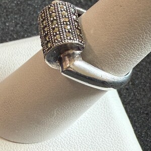 May include: A silver ring with a rectangular top encrusted with small, round, dark-colored stones. The ring is on a finger, showcasing its design and craftsmanship. The ring's band is smooth and polished.