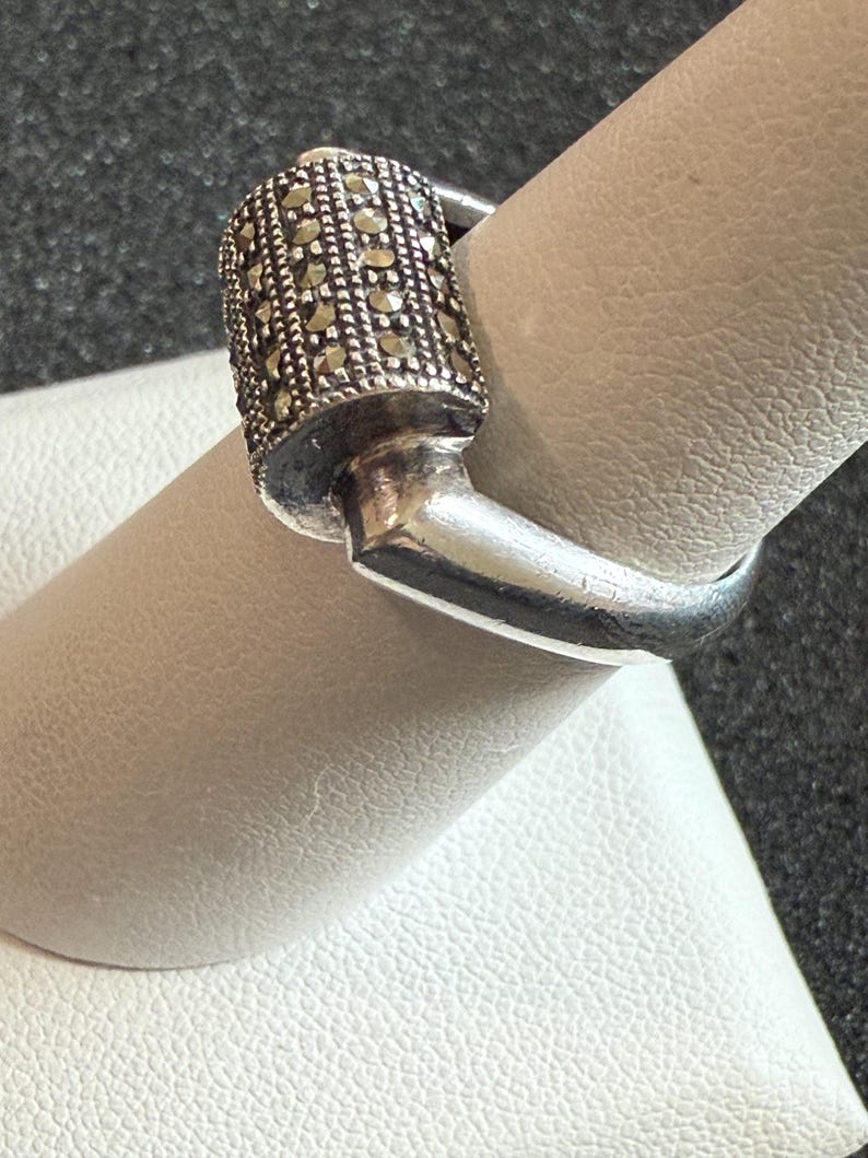 May include: A silver ring with a rectangular top encrusted with small, round, dark-colored stones. The ring is on a finger, showcasing its design and craftsmanship. The ring's band is smooth and polished.