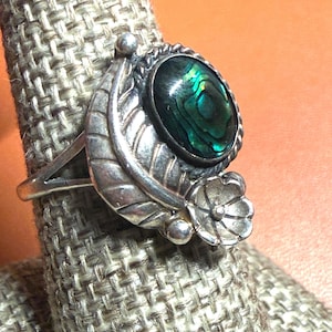 May include: A silver ring featuring an oval abalone shell gemstone with iridescent green and blue hues. The ring has a leaf and flower design on the band, adding a nature-inspired touch to the jewelry.