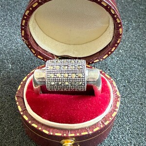 May include: A silver ring with a cylindrical center section encrusted with small, square-cut stones. The ring is displayed inside an open, round, burgundy jewelry box with a red velvet interior and gold-colored trim.