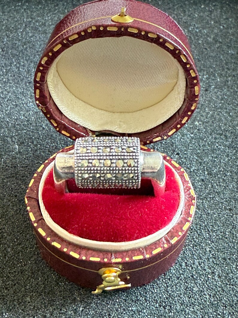 May include: A silver ring with a cylindrical center section encrusted with small, square-cut stones. The ring is displayed inside an open, round, burgundy jewelry box with a red velvet interior and gold-colored trim.